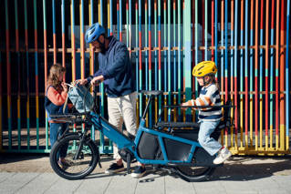 Father and two children make use of an electric cargo bike with space to carry luggage and let small children ride.