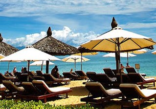 Dense sets of luxury reclining sunbeds are lined close together under parasols on a golden sand beach with turquoise sea.