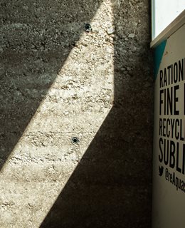 An internal photograph of a Brighton Waste House Wall created from 10 tonnes of rammed chalk. Textured grey wall with sunlight and oblique view of signage.