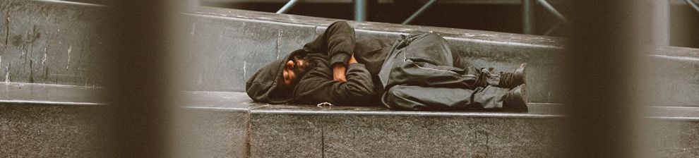 Photographed through railings, a homeless person sleeps on the steps of a modern building. Courtesy Jon Tyson unsplash.