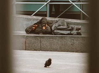 Photographed through railings, a homeless person sleeps on the steps of a modern building. A pigeon on the square in front. Courtesy Jon Tyson unsplash.