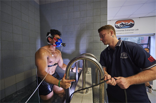 Bare chested male athlete emerges from a pool with monitoring devices, observed by researcher. Image from the Environmental Extreme Lab at the University of Brighton.