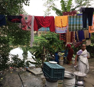 An outdoor river dwelling scene in India with a water pump and plastic bottles. Laundry hangs from a line above two women working.