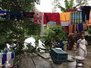 An outdoor river dwelling scene in India with a water pump and plastic bottles. Laundry hangs from a line above two women working.
