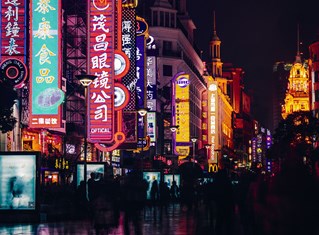 Street scene in Shanghai, China, showing bright neon worded signs down a dark pedestrianised avenue. Courtesy of Krzysztof Kotkowicz and Unsplash.