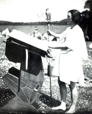 Outdoor photograph in grainy black and white. Kinsperson stands at a carved lectern dressed in a white robe holding an carved falcon staff. Image used through Annebella Pollen's cultural aesthetic history research courtesy of Kibbo Kift Foundation and Don