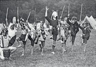 Black and white photograph, 1928. Line of boys and one central adult male all wearing elaborate head-dresses, carrying staffs, some with shields, otherwise rustic shorts and jerkins. Right legs raised in ritual dance or pose. Adult wears large 'White Fox'