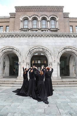 Alice Fox directs performers outside gallery at Korean Disability Arts and Culture festival. Outdoor photograph with gallery building with archways in background. Group of artists dressed in black stand with arms raised.