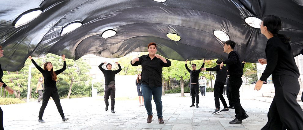 Performance directed by Alice Fox at Korean Disability Arts Festival. Vast black fabric square billows into the air above a group of artist participants, each of whom now moves inwards, under it.