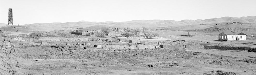 Black and white photography of desert landscape with distant hills. To the far left a tower structure, to the far right a ruined building, evidence of mined land dominates the image. Research photography by Xavier Ribas for Traces of Nitrate, Desert Trail