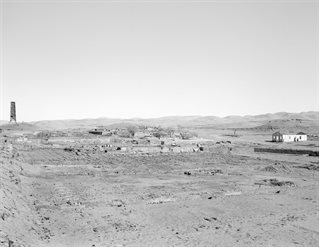 Black and white photography of desert landscape with distant hills. To the far left a tower structure, to the far right a ruined building, evidence of mined land dominates the image. Research photography by Xavier Ribas for Traces of Nitrate, Desert Trail