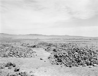 Black and white photograph of desert landscape with distant mountains and, in foreground, rubble and boulders from mining industry. Xavier RIbas research photography for Traces of Nitrate, Desert Trails.