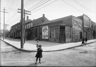 Black and white photograph taken 1912 of a street corner with a wide pavement and telegraph posts on the left. Two small girls are captured in the foreground and two small boys in the background. Arthur Goss, New  Registry Office Site, May 15, 1912 