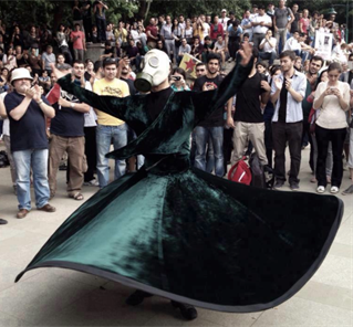 A crowd in contemporary park setting watch a performer with a wide spread skirt in green velvet. A Whirling Dervish.