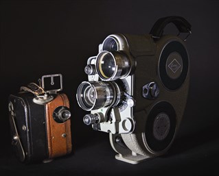 Two antique film cameras, artistically photographed against dark background.One large chrome example is a Eumig 16mm amateur cine camera from the 1950s. The other a Pathé Baby 9.5mm amateur cine camera from the 1920s.