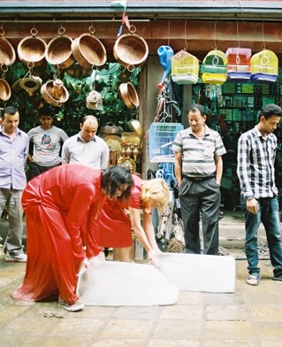 A street scene in Nepal. Two artists, Alice Fox and Ashmina Ranjit, each push a large block of ice along the road outside a shop with onlookers. Performance of Paradox of Praxis 1 #2 (Pushing Together)