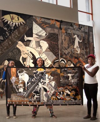 A vast tapestry remaking of the iconic Guernica with locally created differences. In the foreground three craftspeople hold up a smaller version of a woven Guernica piece. Research by Nicola Ashmore with craftspeople from Keiskamma, South Africa_Guern