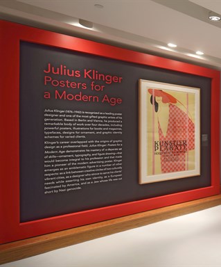 Internal exhibition space with red and dark grey livery on white floor. Title banner reads Julius Klinger, Posters for a Modern Age