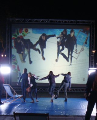 Darkened outdoor space with installation of large screen and blue ground mat. People dance and play on the mat, viewing themselves on the screen.