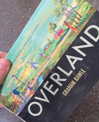 Hand holding Graham Rawle's overland, rotating the cover to see the horizontal format. Large title, Overland. Cover half half between 1950s colourful poster art landscape and black and white industrial image.