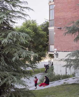 Photograph by Xavier Ribas. Contemporary brick building, pine tree in foreground with isolated figures on slope beyond near grass. Trinitat Nova Barcelona.