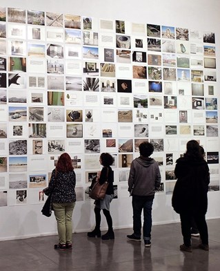 Exhibition installation photograph of Trafficking the Earth with silhouette figures staring up at the many wall mounted photographs.