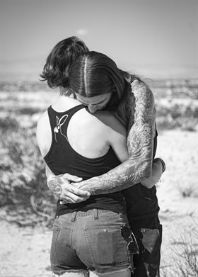 Black and white image of a couple hugging in a desert landscape. Male figure leans over female with tattooed arms clasped around her. Zoe Childerley, Dinosaur Dust.