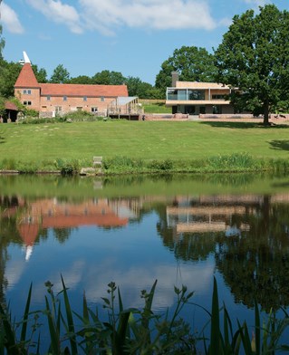 Landscape with large pond in foreground, lawn and blue sky. Traditional oast house and farm building to the left. To right two story contemporary structure with large floor to ceiling windows, flat roof and full length horizontal balcony, partially hidden