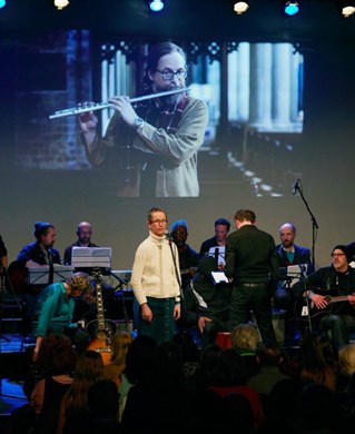 Orchestra in darkened space performing in front of a screen projection of flute player.