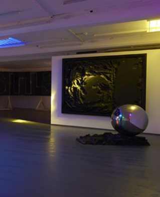 Indoor exhibition space with sculptures. A curved blackboard, wall mounted blackboard and silver sphere with black ribbon.