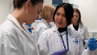 Post-doctoral chemistry researcher Dr Wulan Koagouw in laboratory with students. Scientists in white coats with blue gloves and equipment.