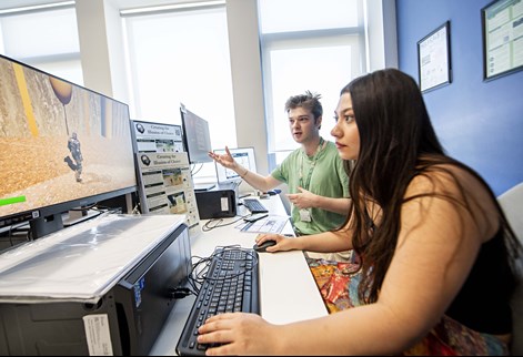 Two students in a computer suite playing a 3D game on a large monitor.