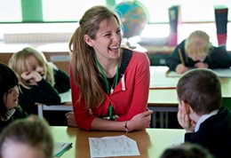 Primary school teacher at a desk laughing with the children around her