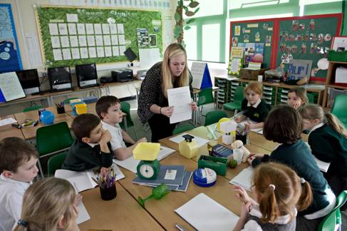 Primary school classroom teacher stands at a table showing a paper-based resource. Ten young primary learners sit around the table eagerly watching.