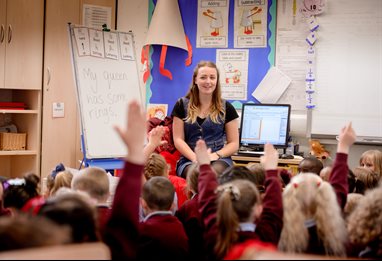 School teacher in front of her pupils, all with their hands up to answer a question