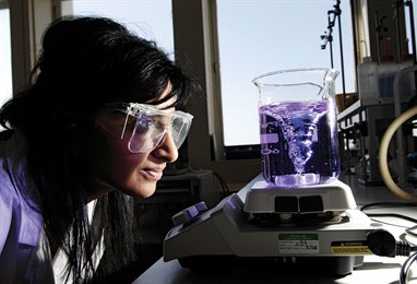 Student in a lab coat and safety specs watching a purple liquid rotate in a glass beaker
