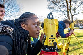 Construction female student looking through total station surveying equipment outside