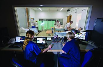 Two apprentices looking at a mock patient ward from behind a screen.