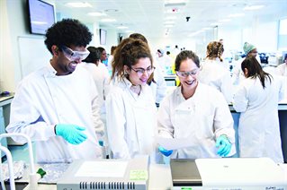 Three students at a lab bench wearing white coats and protective goggles