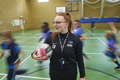 Smiling student gym teacher holding football in gym with children running around them