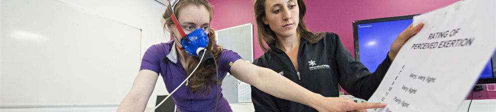 Image of Brighton University sports science testing shows two researchers one with a mask on an exercise machine, the other holding a large paper reading Rating of Perceived Exertion to which the exerciser points for Fairly Light.