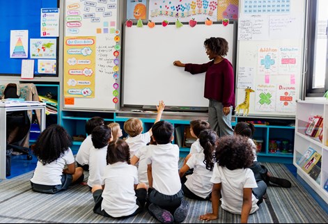 Female teacher leading a whole-class lesson with primary pupils participating in classroom discussion.