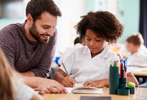 Male primary teacher supporting a pupil with reading and writing during a classroom activity.
