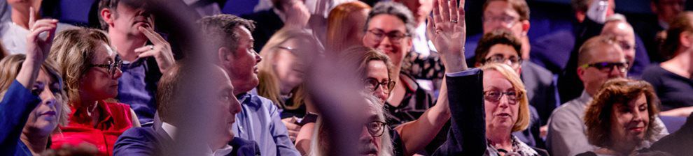 An audience of people, some with hands raised to ask a question