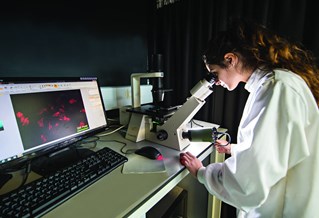 Biomedical science researcher in white coat using an electronic microscope with visual feed to nearby computer screen