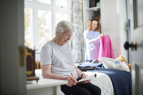 Elderly woman sitting on a bed with a care worker in the background