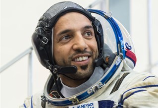 Emirati astronaut Sultan Al Neyadi with grey white space suit with UAE flag, helmet removed. He sits in front of the Soyuz spacecraft simulator in 2019.