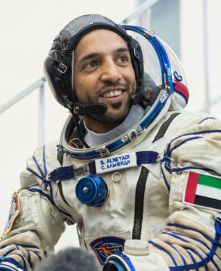 Emirati astronaut Sultan Al Neyadi with grey white space suit with UAE flag, helmet removed. He sits in front of the Soyuz spacecraft simulator in 2019.