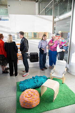 A beanbag, chair, and table on top of a green mat made of fake grass surrounded by people.