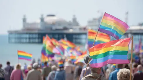 A crowd waves LGBTQ+ Pride flags in front of Brighton Pier